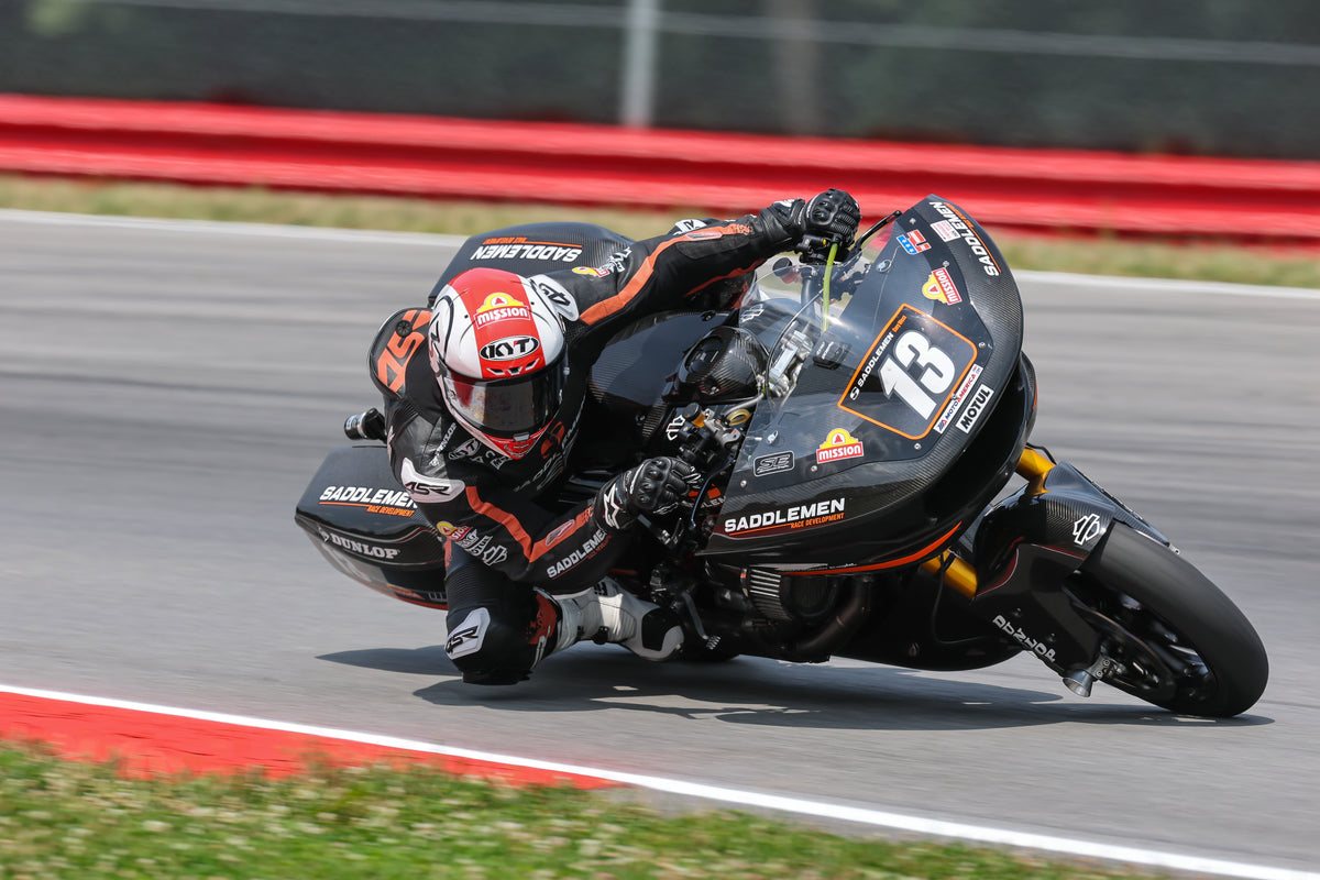 Cory West leans sharply into a turn on a racetrack, wearing a black, red, and white racing suit and his custom KYT KX-1 Race GP Motorcycle Helmet while riding a black number 13 saddlemen Harley-Davidson