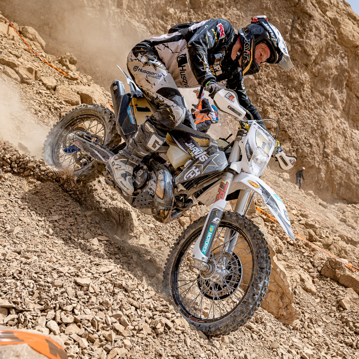 Graham Jarvis in full protective gear leans a white and black Husqvarna dirt bike over a rocky, dusty incline.