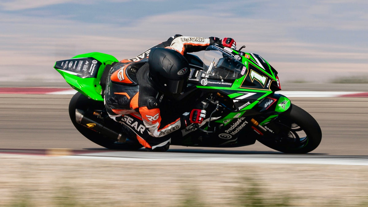 A green and black Kawasaki motorcycle with racing decals is ridden by Anthony Norton in a black and orange suit on a blurred racetrack.
