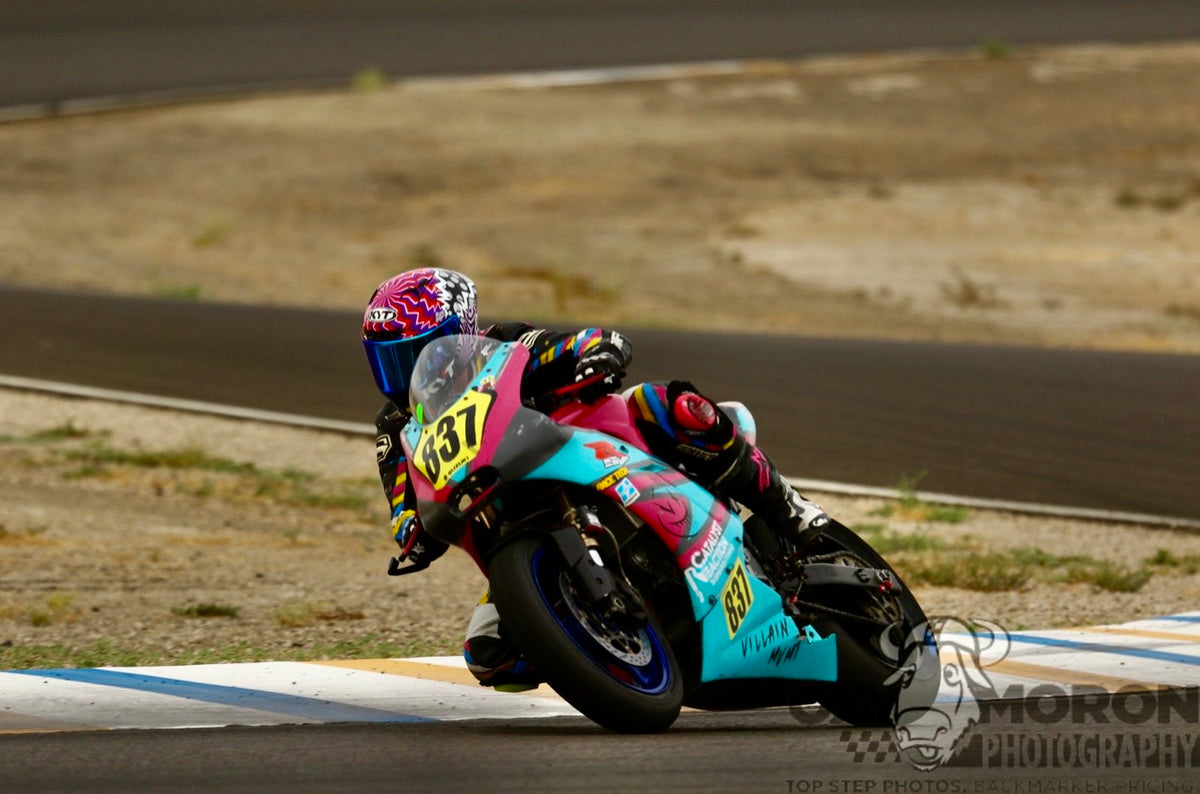 A racer leans sharply into a turn on a turquoise, pink, and black motorcycle on a racetrack, with dry grass and asphalt in the background.
