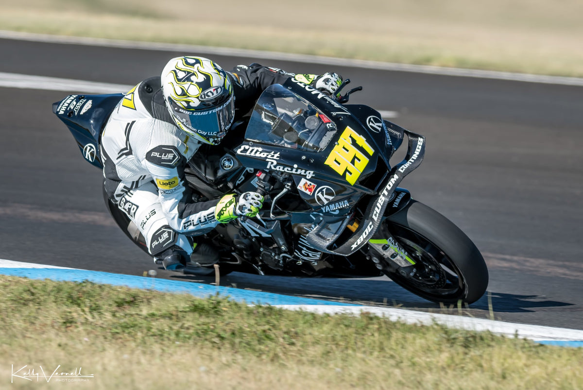 Kreece Elliot leans sharply into a turn on a racetrack, wearing a white, black, and yellow racing suit and KYT KX-1 Race GP Motorcycle Helmet with a black racing bike featuring the number 88 and sponsor logos.